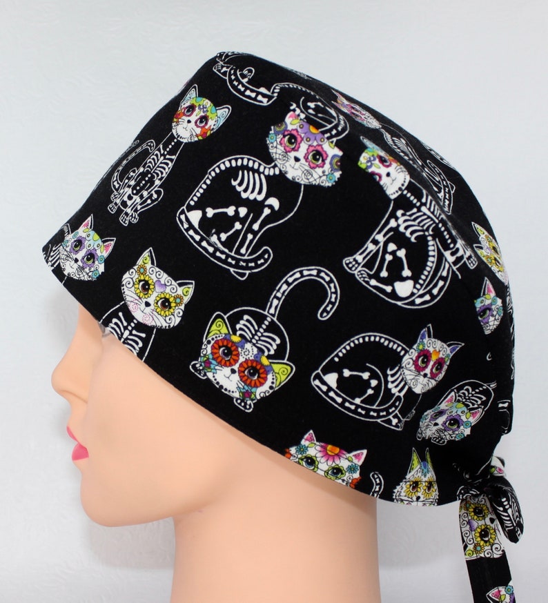 Puede incluir:  gorro de quir&oacute;fano negro con un estampado de gato colorido del D&iacute;a de Muertos. Los gatos tienen calaveras de az&uacute;car y dise&ntilde;os florales.