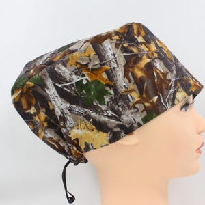 May include: A camouflage print scrub cap with a drawstring closure. The cap features a pattern of brown, green, and yellow leaves and branches. The text "ADVANTAGE" is visible on the side of the cap.