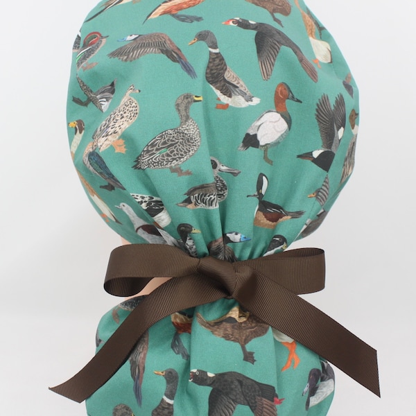 May include: Teal blue fabric scrub hat with a brown ribbon tied around it. The fabric has a pattern of various types of ducks in different colours.
