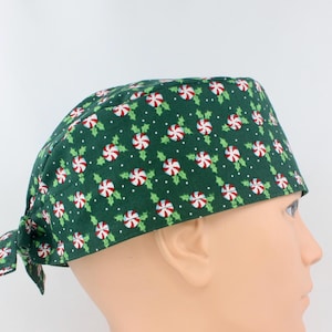 May include: A dark green scrub cap featuring a repeating pattern of red and white peppermint sweets, green holly leaves, and small white dots. The cap has a tie-back closure at the nape of the neck and a structured band that fits around the head.