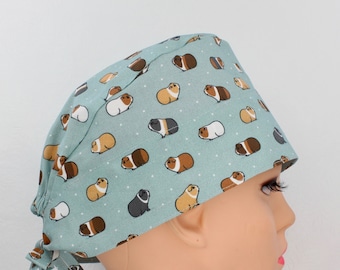 May include: A light teal surgical scrub cap featuring a repeating pattern of cartoon guinea pigs in various colors. The guinea pigs are brown, white, black, and tan. The cap has a tie closure at the back and small white dots scattered across the fabric.