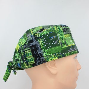 May include: A green and black surgical scrub cap featuring a circuit board design. The cap is tied at the back and is made of a lightweight fabric. The design includes various electronic components and pathways.