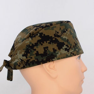 May include: A camouflage patterned scrub cap in shades of green, brown, and black. The cap is tied at the back and is shown on a mannequin head. The fabric has a pixelated design.