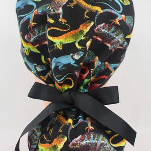 May include: A black scrub cap featuring a colorful lizard pattern. The lizards are in shades of green, yellow, orange, blue, and purple. A black ribbon is tied in a bow at the back of the cap. This cap is designed for medical professionals.