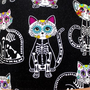Puede incluir: Tela negra con un patr&oacute;n repetido de gatos esquel&eacute;ticos blancos con caras de calavera de az&uacute;car de colores. Los gatos tienen detalles rosas, azules, verdes y amarillos.