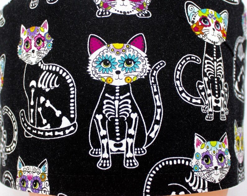Puede incluir: Tela negra con un patr&oacute;n repetido de gatos esquel&eacute;ticos blancos con caras de calavera de az&uacute;car de colores. Los gatos tienen detalles rosas, azules, verdes y amarillos.