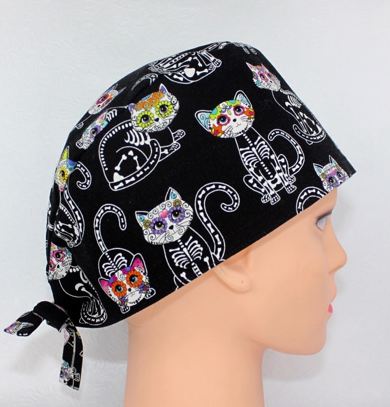 Puede incluir:  gorro de quir&oacute;fano negro con un estampado de gato del D&iacute;a de los Muertos colorido. Los gatos tienen esqueletos blancos y flores coloridas en sus caras.