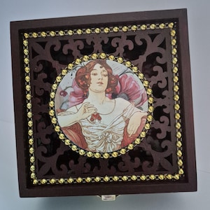 May include: A dark brown wooden jewellery box with a decorative cutout design and a gold beaded border. The lid features a circular image of a woman with red hair and a floral background. The woman is holding a red heart-shaped object.