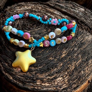 Puede incluir: Una pulsera de cuentas hecha a mano con las palabras "OFU STAR" deletreadas con cuentas de letras coloridas. La pulsera presenta un dije de estrella dorada y est&aacute; adornada con cuentas azules, rosas y marrones. Se muestra sobre una superficie de madera oscura.