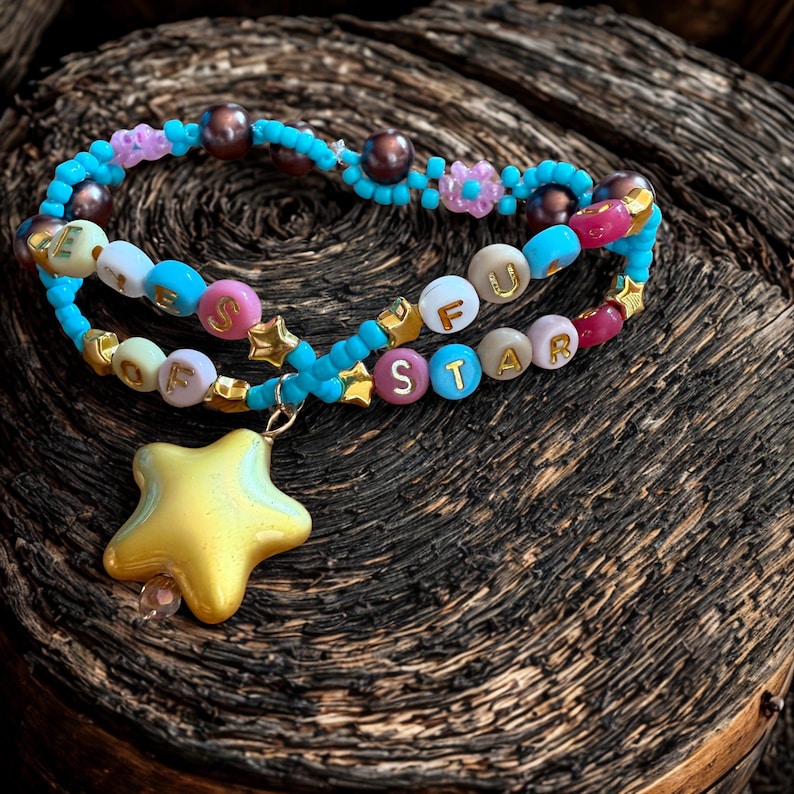 Puede incluir: Una pulsera de cuentas hecha a mano con las palabras "OFU STAR" deletreadas con cuentas de letras coloridas. La pulsera presenta un dije de estrella dorada y est&aacute; adornada con cuentas azules, rosas y marrones. Se muestra sobre una superficie de madera oscura.