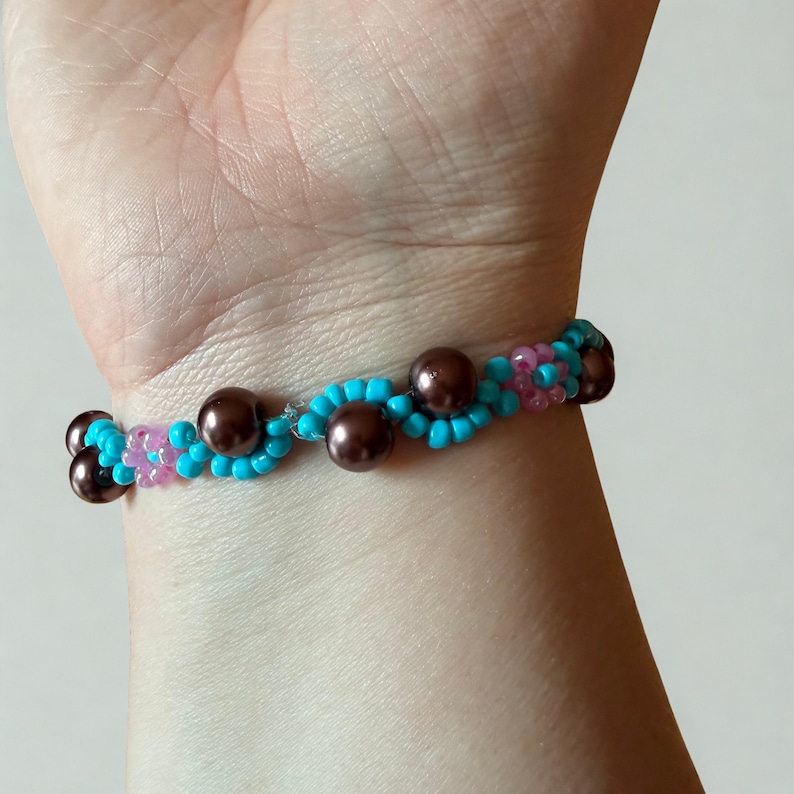 Puede incluir: Una pulsera de cuentas con un patr&oacute;n de ondas. La pulsera presenta cuentas turquesas y rosas, con cuentas marrones con forma de perla intercaladas. La pulsera se lleva en la mu&ntilde;eca.
