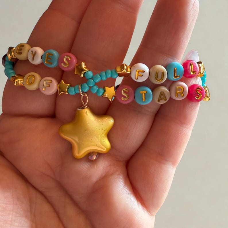 Puede incluir: Dos pulseras de cuentas con las palabras "EYES OF" y "FULL STARS" deletreadas en cuentas de letras coloridas. Las pulseras est&aacute;n conectadas por cuentas turquesas y dijes de estrella dorada, con un gran colgante de estrella dorada.