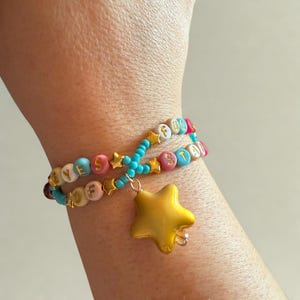 Puede incluir: Dos pulseras de cuentas con las palabras "YES" y "FOOL" y "STAR" deletreadas en cuentas de letras coloridas. Las pulseras est&aacute;n conectadas por cuentas turquesas y dijes de estrella dorada, con un gran colgante de estrella dorada.