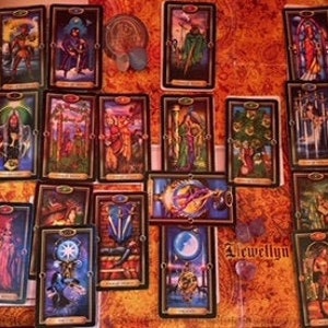May include: A spread of tarot cards featuring various illustrations and symbols. The cards are arranged on a wooden surface, with some cards face up and others face down. The colors are rich and vibrant, with gold and jewel tones.