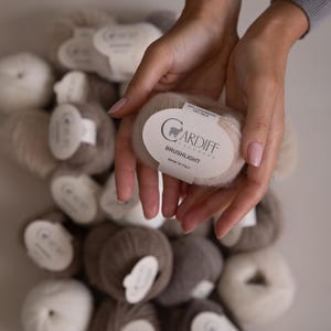 Peut inclure: Une collection de pelotes de laine douces et duveteuses dans des tons beige, crème et marron. Chaque pelote porte une étiquette en papier blanc avec le texte Cardiff Cashmere, Brushlight, Made in Italy, 82% Cashmere et 18% Silk imprimé.