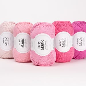 Pode incluir: Cinco novelos de fio de algodão dispostos em fila, exibindo um gradiente de tons de rosa, desde o blush pálido até ao magenta profundo. Cada novelo apresenta uma etiqueta de papel branco com o texto preto "DROPS Magic Uni Colour Classic 8/4 cotton".