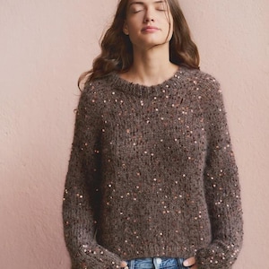 Peut inclure: Un pull en tricot à manches longues marron doté d'une encolure ronde et de paillettes métalliques éparpillées sur tout le tissu. Le pull présente un aspect duveteux et texturé, associé à un jean bleu avec un bouton en métal visible.