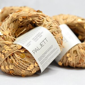 May include: Two skeins of beige yarn intertwined with small, reflective gold sequins. Each skein features a white paper label with the text Sandnes Garn, Den Norske Garnfabrikken Siden 1888, and Paljett, along with product details in small print.