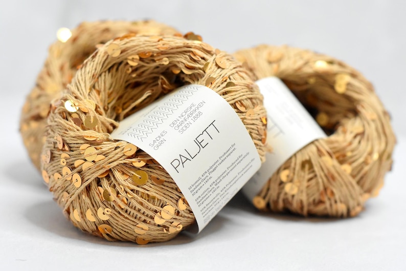 May include: Two skeins of beige yarn intertwined with small, reflective gold sequins. Each skein features a white paper label with the text Sandnes Garn, Den Norske Garnfabrikken Siden 1888, and Paljett, along with product details in small print.