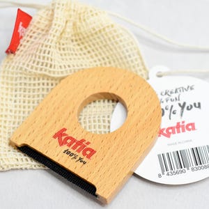 May include: A wooden lint remover with a handle and a mesh bag. The lint remover has a light wood finish with the Katia logo in red. A white tag with the Katia logo and text is also present.