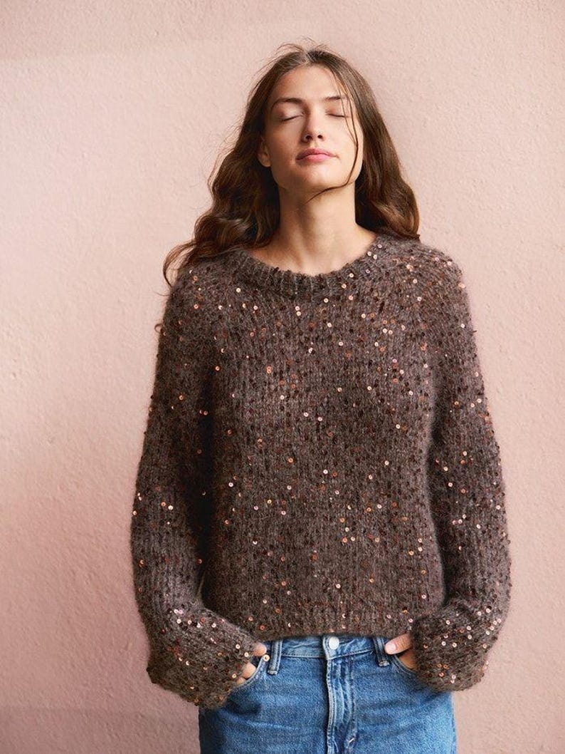 May include: A brown long-sleeve knit sweater featuring a crew neckline and scattered copper-colored sequins throughout the fabric. The sweater has a fuzzy, textured appearance and is paired with blue denim jeans, with hands tucked into the pockets.