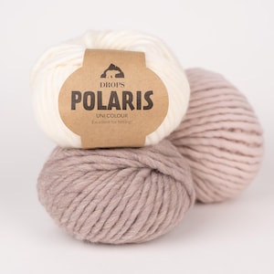Peut inclure: Trois pelotes de laine épaisse et volumineuse empilées. La pelote du haut est blanc cassé, tandis que les deux du bas sont d'une teinte mauve-brun. Une étiquette en papier brun entoure la pelote supérieure avec le texte DROPS POLARIS UNI COLOUR.