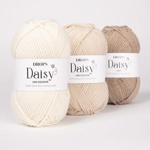 Könnte beinhalten: Drei Knäuel Drops Daisy Garn in einer Reihe angeordnet. Das erste ist cremeweiß, das zweite hellbeige und das dritte in einem dunkleren Beigeton. Jedes Knäuel hat ein weißes Papieretikett mit dem Text Drops Daisy, Uni Colour, 100% extra fine merino wool.