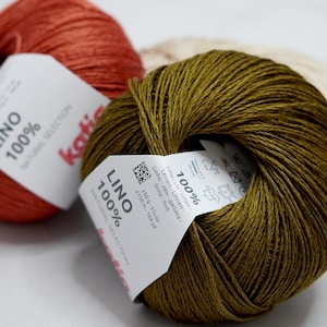Può includere: Primo piano di tre gomitoli di filato Katia Lino 100% nei toni ruggine, verde oliva e crema. Ogni gomitolo ha un'etichetta bianca con il nome del marchio e i dettagli del prodotto. Il filato è 100% lino, con 150g e 150 metri.