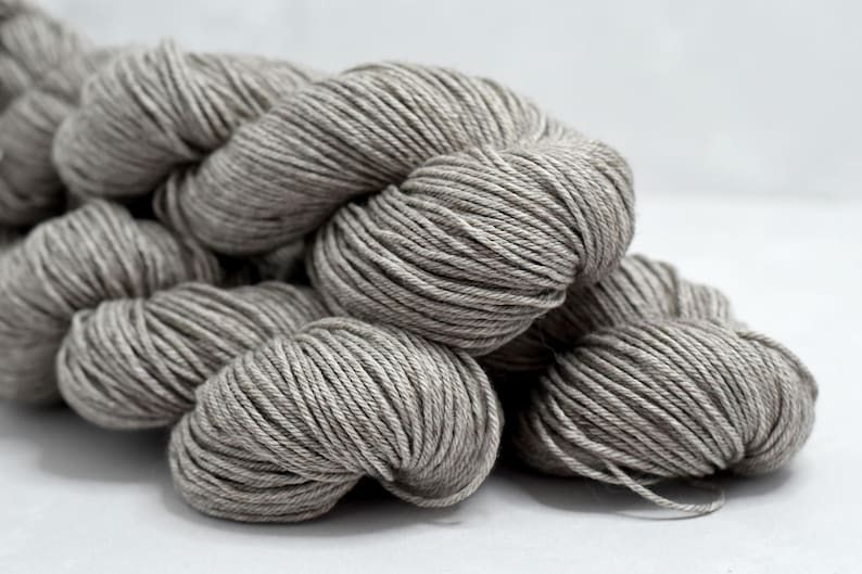 May include: Close-up of several skeins of gray yarn. The yarn is tightly wound into round bundles, with visible strands and a soft, textured appearance. The neutral color suggests versatility for various knitting or crochet projects.