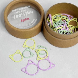 May include: A round cardboard container filled with colourful cat-shaped stitch markers. The container has a clear lid with the text "100% Creative 100% Fun 100% You katia Cat Stitch Markers".