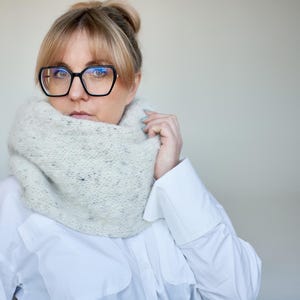 May include: A woman wearing a light gray knit cowl neck scarf. The scarf has a speckled pattern. She is wearing a white button-down shirt and large black-framed glasses. Her hair is pulled back in a bun.