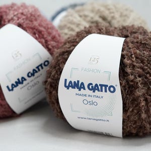 Può includere: Primo piano di tre gomitoli di lana Lana Gatto nei toni del marrone, beige e malva. Ogni gomitolo ha un'etichetta con la scritta "Lana Gatto", "Made in Italy" e "Oslo". La lana ha un aspetto strutturato e a ricciolo.