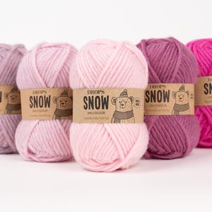 Könnte beinhalten: Eine Reihe von fünf Garnknäueln der Marke Drops Snow in verschiedenen Rosa- und Violetttönen. Jedes Knäuel hat eine beigefarbene Papierbanderole mit dem Text Drops Snow, Uni Colour, einer Bärenillustration mit Mütze und Schal sowie 100% wool.