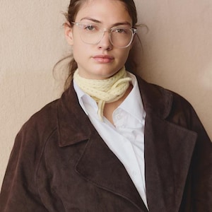 May include: A dark brown suede coat worn over a crisp white collared button-down shirt. A pale yellow knit neck scarf is tied around the neck. The look is completed with clear-framed eyeglasses featuring a rounded shape and silver hardware accents.