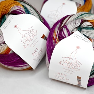 Puede incluir: Dos ovillos de lana de colores con etiquetas blancas que dicen "Laines du Nord" y "Art Sock". La lana es una mezcla de naranja, morado, verde y blanco.