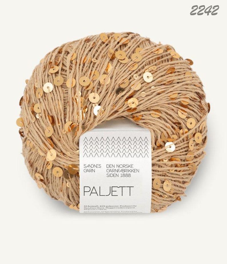 May include: A round ball of beige yarn embellished with small, reflective gold sequins. A white paper label wraps around the center, featuring the text Sandnes Garn, Den Norske Garnfabrikken Siden 1888, Paljett, and the number 2242 at the top.