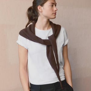Könnte beinhalten: Ein schlichtes weißes Kurzarm-T-Shirt kombiniert mit einem dunkelbraunen Strickpullover, der über die Schultern gelegt und vorne verknotet ist, getragen zu einer schwarzen Hose. Das Ensemble besticht durch eine schlichte Optik in neutralen Farben.