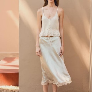 May include: A cream-colored sleeveless camisole top with thin straps, a button-down front, and a scalloped lace hem, paired with a light champagne-colored midi-length satin skirt featuring a delicate lace trim along the bottom hemline.