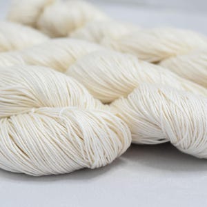 Peut inclure: Gros plan de plusieurs écheveaux de fil blanc cassé. Le fil est étroitement enroulé, avec des brins visibles et un aspect doux et texturé. Les écheveaux sont disposés en chevauchement, suggérant un produit artisanal ou textile.