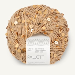 Può includere: Un gomitolo di filato beige decorato con piccole paillettes rotonde color oro metallizzato. Un'etichetta di carta bianca avvolge il centro con il testo Sandnes Garn, Den Norske Garnfabrikken Siden 1888 e Paljett, oltre alle specifiche tecniche.