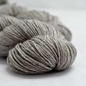 May include: Close-up of a skein of gray yarn. The yarn is tightly wound, showing the individual strands. The color is a neutral gray, suitable for various knitting or crochet projects. The yarn is on a white surface.
