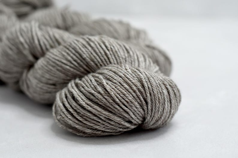 May include: Close-up of a skein of gray yarn. The yarn is tightly wound, showing the individual strands. The color is a neutral gray, suitable for various knitting or crochet projects. The yarn is on a white surface.