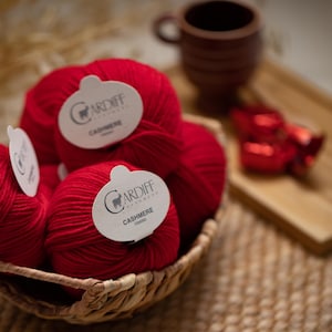 May include: A woven basket filled with several balls of bright red yarn. Each ball features a white paper label with the text Cardiff Cashmere printed in black. The yarn is soft and textured, arranged in a cluster within the basket.