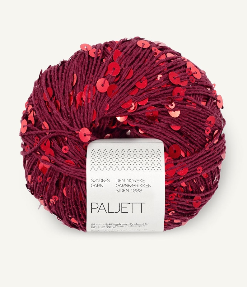 May include: A round ball of deep burgundy yarn embellished with numerous small, shiny red sequins. A white paper label is wrapped around the center, featuring the text Sandnes Garn, Den Norske Garnfabrikken Siden 1888, and Paljett.