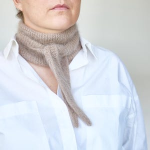 May include: A beige knitted scarf is draped around the neck, with the ends hanging down. The scarf is worn over a white button-down shirt. The scarf appears to be made of a soft, fuzzy material.