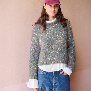 May include: A maroon baseball cap with the text Colorado Rocky Mountain, a grey knit sweater featuring scattered multicolored sequins, a white collared shirt worn underneath, and blue denim jeans. The sweater has a crew neckline and long sleeves.