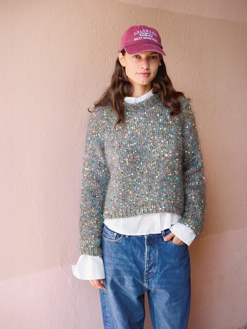 May include: A maroon baseball cap with the text Colorado Rocky Mountain, a grey knit sweater featuring scattered multicolored sequins, a white collared shirt worn underneath, and blue denim jeans. The sweater has a crew neckline and long sleeves.