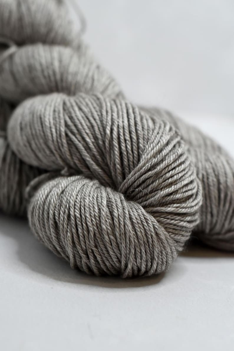 May include: Close-up of several skeins of gray yarn. The yarn is tightly wound, showing individual strands. The color is a neutral gray, suitable for various knitting or crochet projects. The yarn is soft and appears to be made of natural fibers.