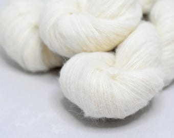 May include: Close-up of several skeins of soft, white yarn. The yarn appears fluffy and textured, with a slight sheen. The skeins are arranged in a loose, organic shape, suggesting a craft or textile project. The background is a plain, light surface.