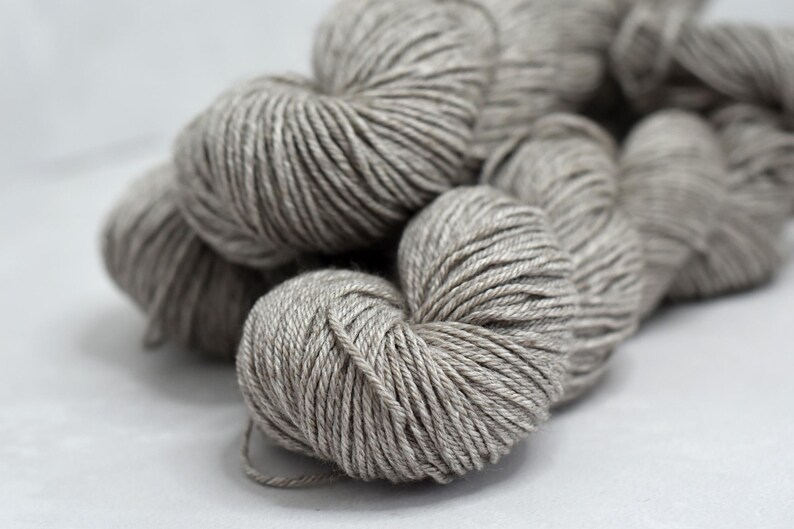 May include: Close-up of several skeins of gray yarn. The yarn is tightly wound into round balls, with visible strands and a soft, textured appearance. The neutral color suggests versatility for various knitting or crochet projects.